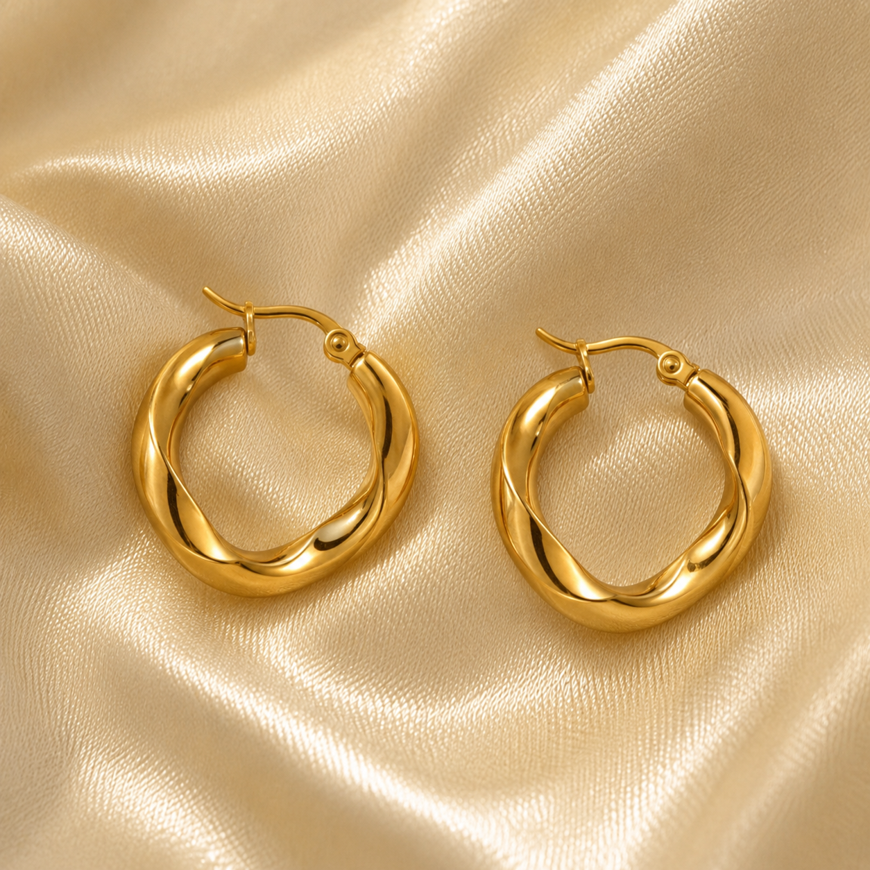 A pair of polished gold hoop earrings with a twisted design, displayed on a draped champagne-colored satin fabric.