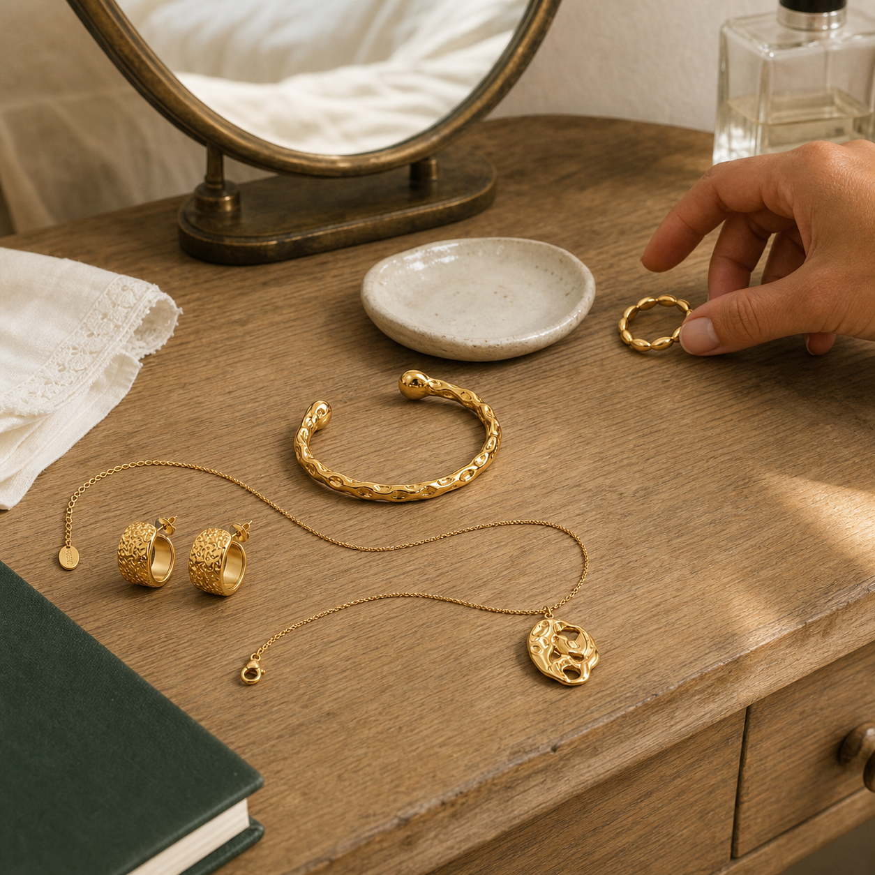 A hand is placing a small gold ring on a wooden table next to a gold bracelet, necklace, and earrings with textured finishes. The scene includes a small mirror, a ceramic dish, a book, and a perfume bottle in a warm, styled setting.