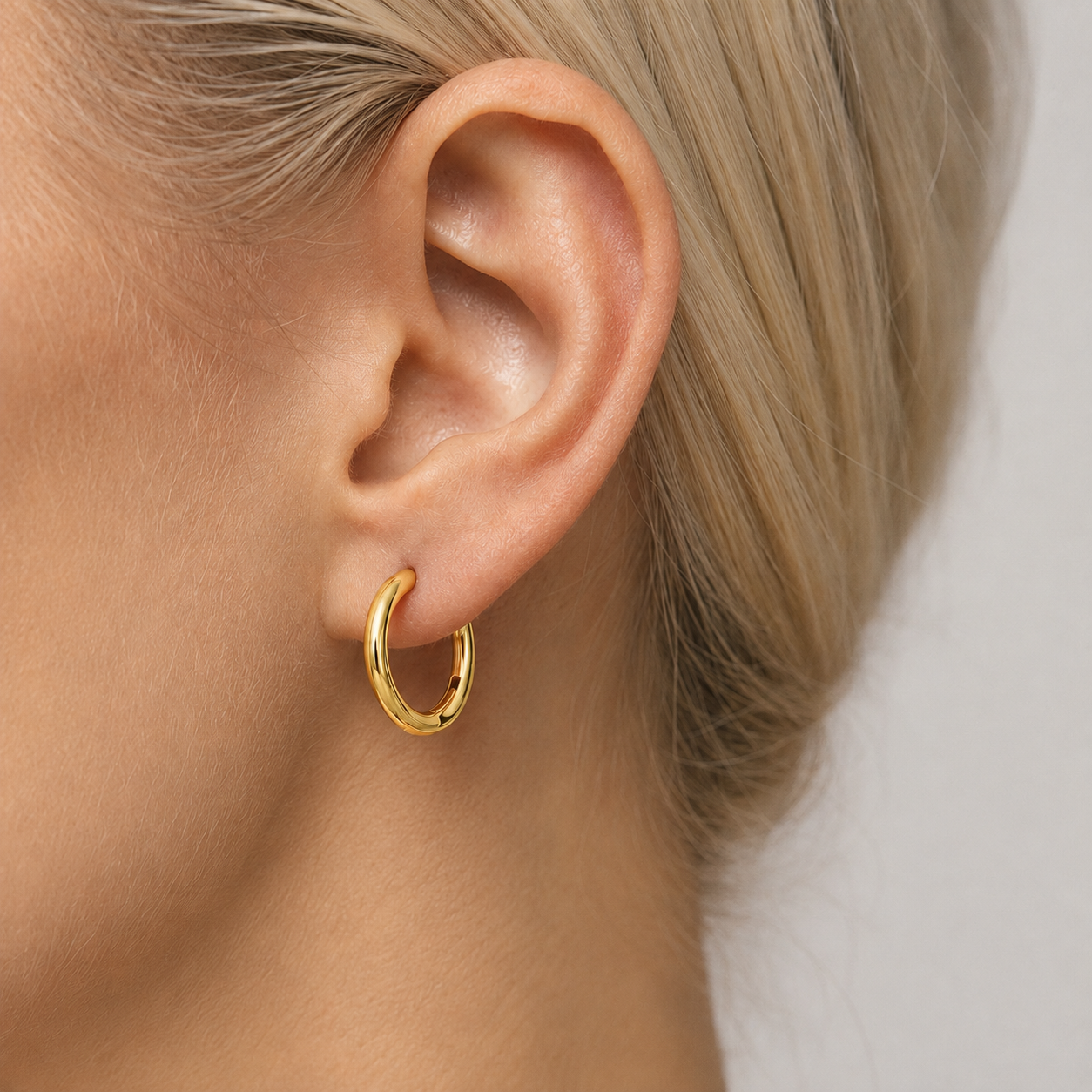 A close-up of a model's ear wearing a small, polished gold hoop earring.