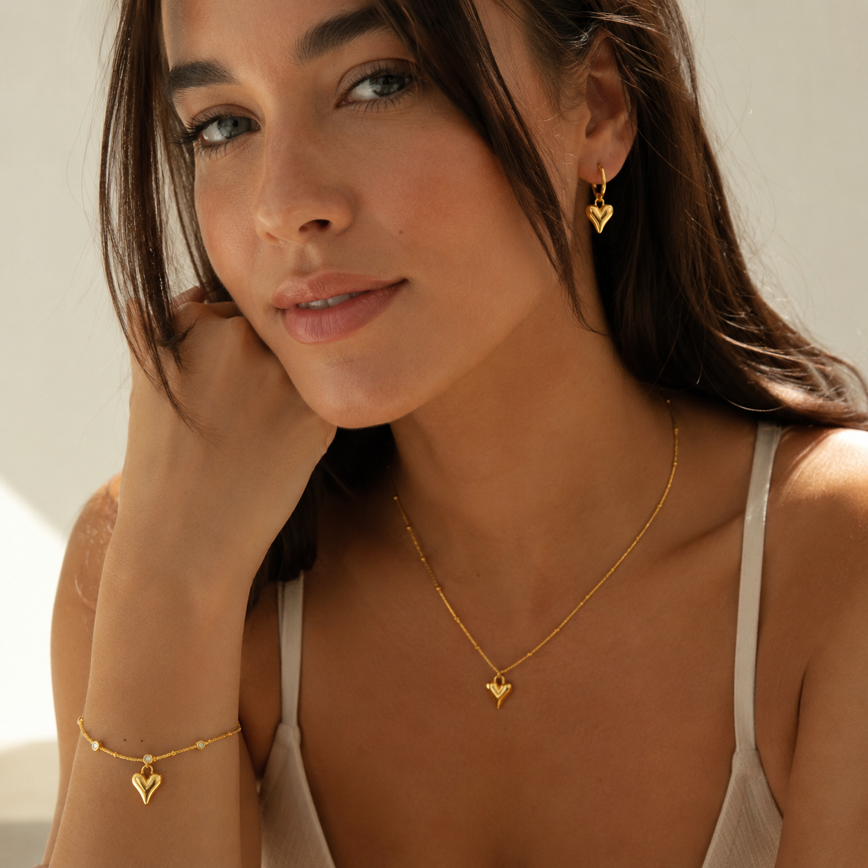 A woman wearing a delicate gold necklace with a small heart pendant, a matching gold bracelet with a heart charm on her wrist, and a small gold heart drop earring.