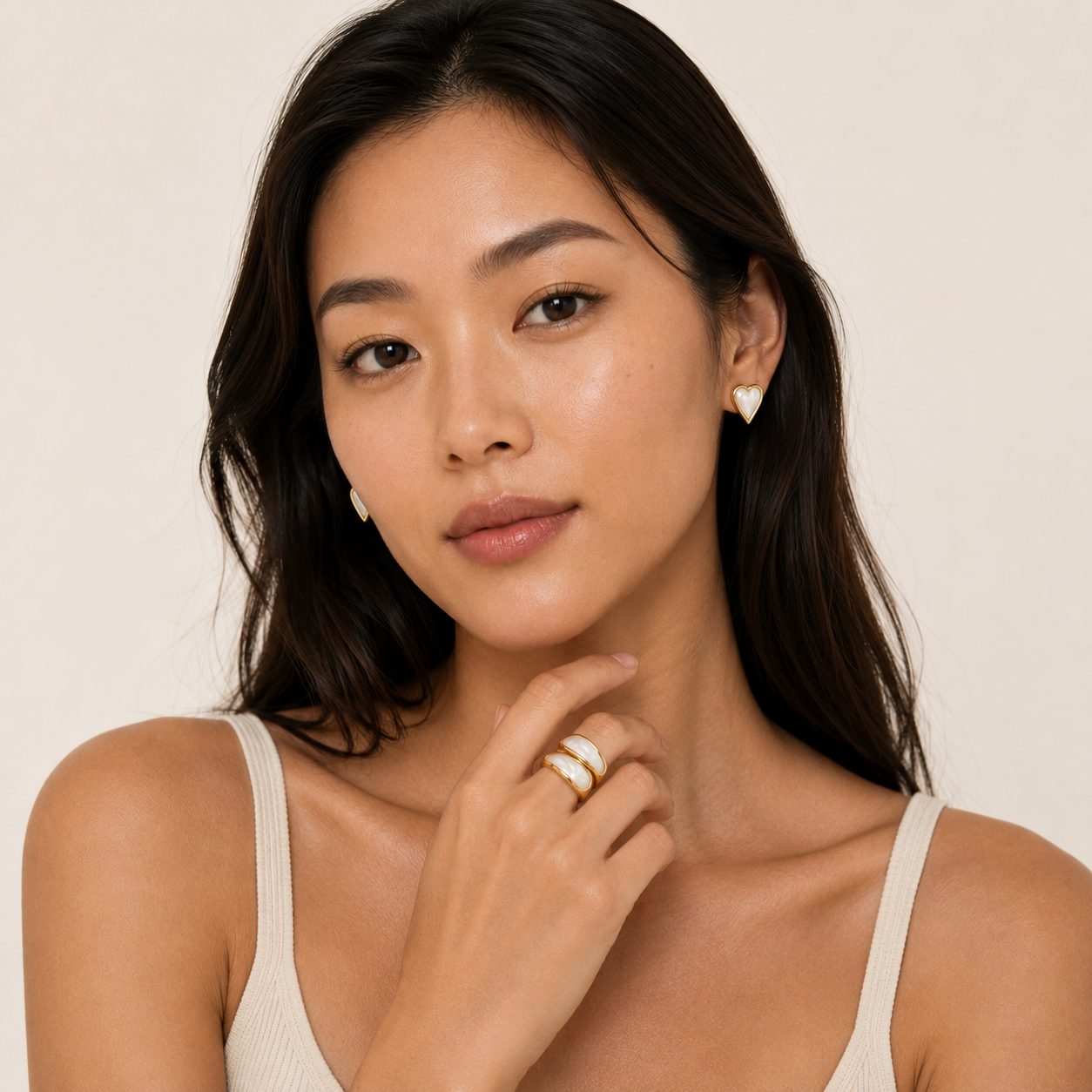 A close-up of a woman wearing a gold ring with a white mother-of-pearl stone on her finger and matching white stud earrings.