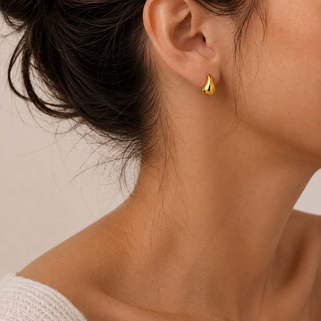 A close-up of a model's ear wearing a small, polished gold teardrop stud earring.