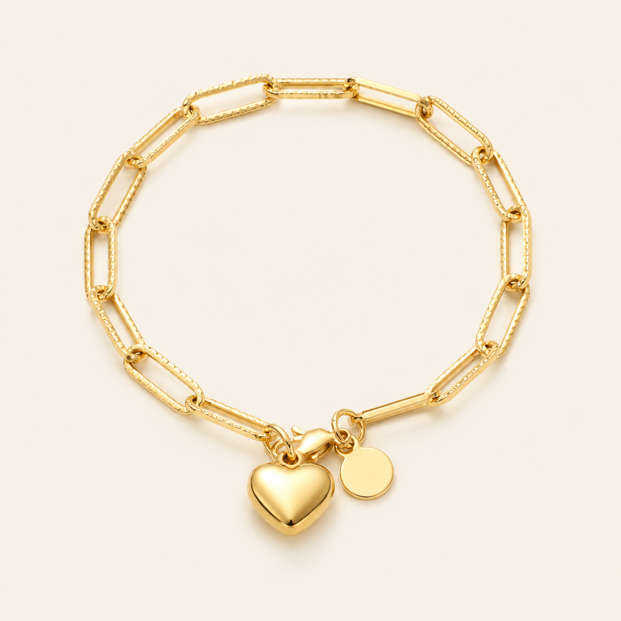 A gold paperclip chain bracelet with a polished heart charm and a small round disc charm.