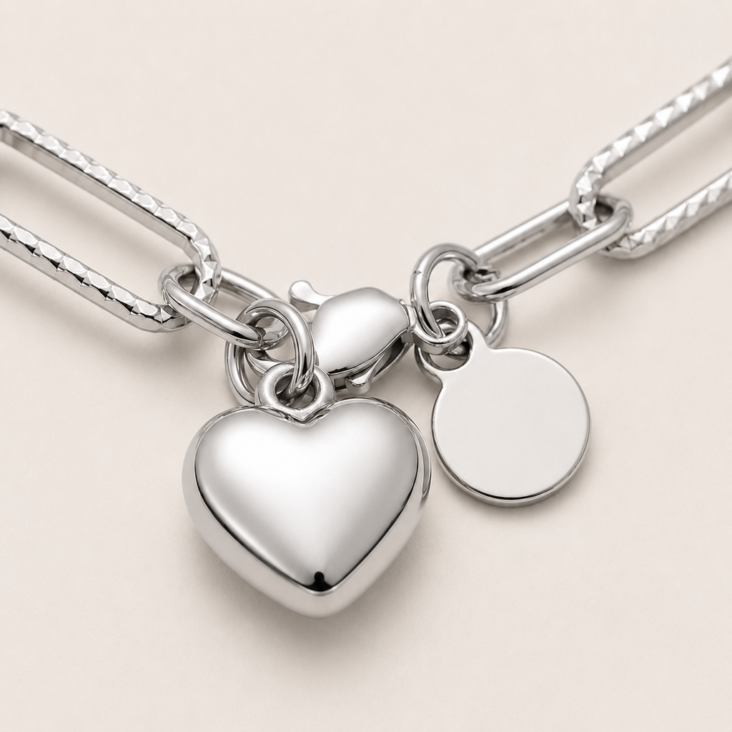 A close-up of a silver chain necklace with a large polished heart charm and a blank round disc tag.