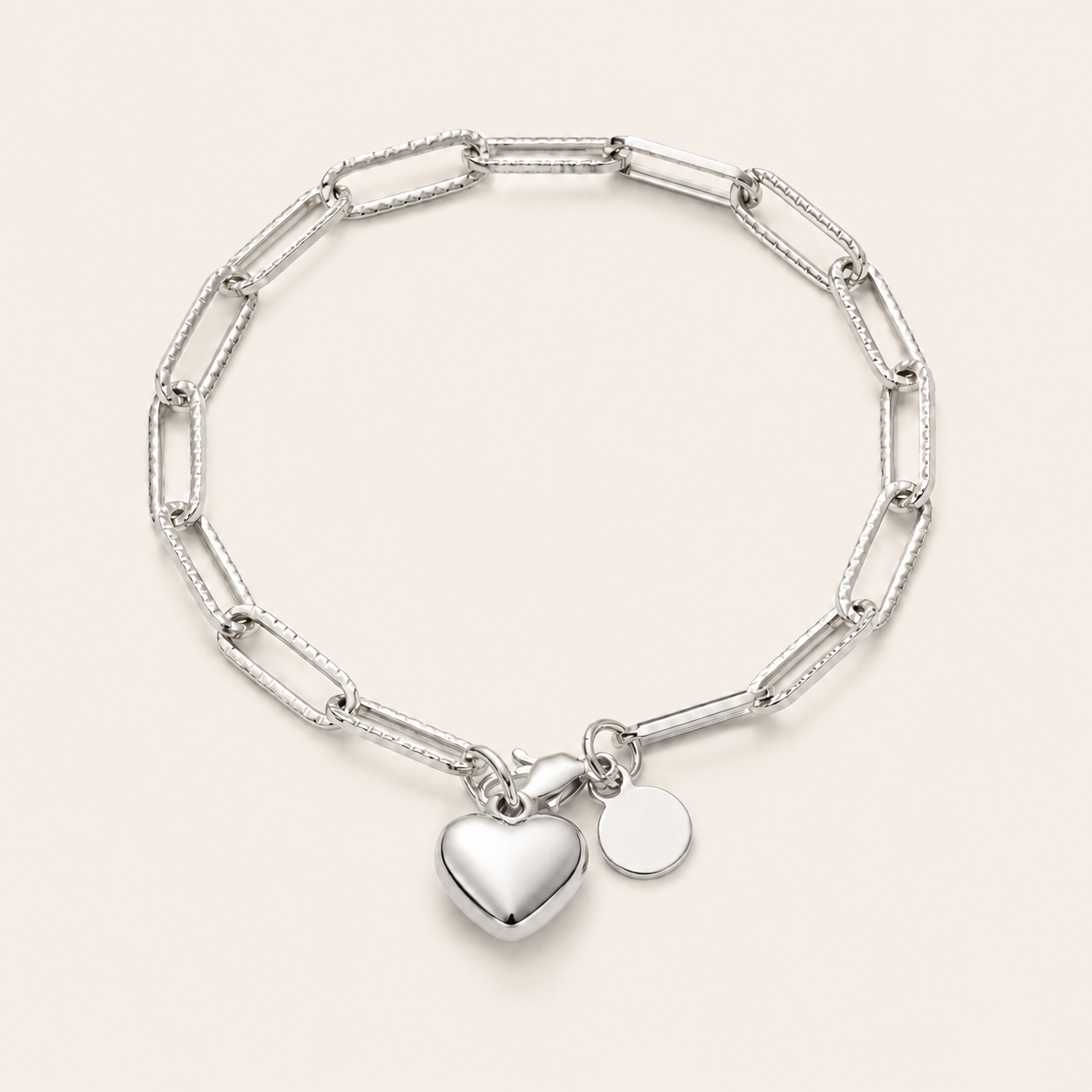 A silver paperclip chain bracelet with a polished heart charm and a small round disc charm.