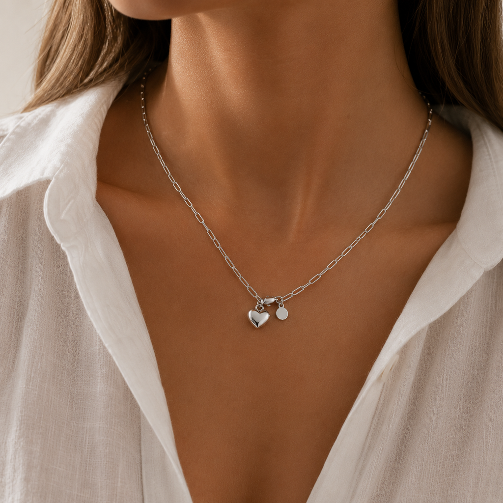 A close-up of a model wearing a delicate silver chain necklace with a small heart charm, styled with a white collared shirt.