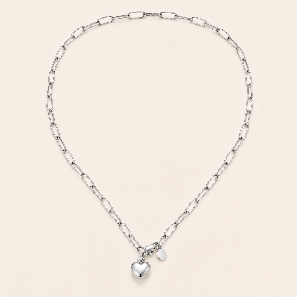 A silver paperclip chain necklace with a small polished heart charm and a tiny oval tag.