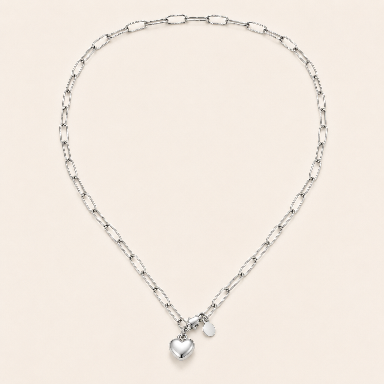 A silver paperclip chain necklace with a small polished heart charm and a tiny oval tag.
