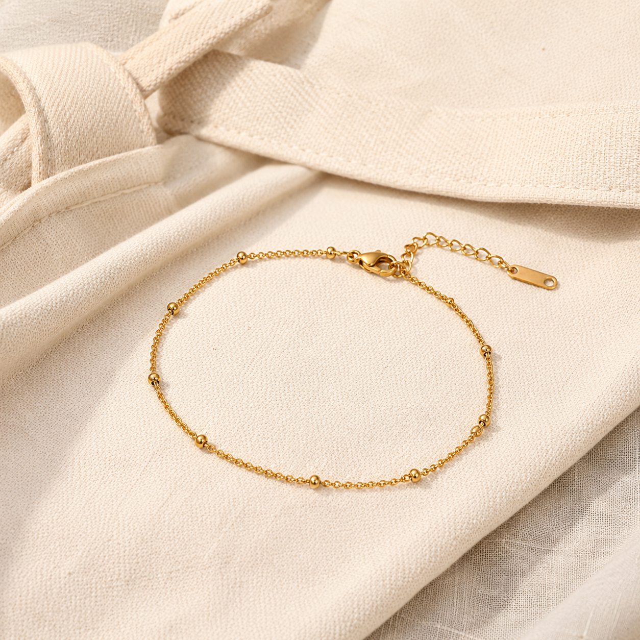 A delicate gold chain anklet with small bead accents and an adjustable clasp, displayed on a soft cream-colored fabric background.