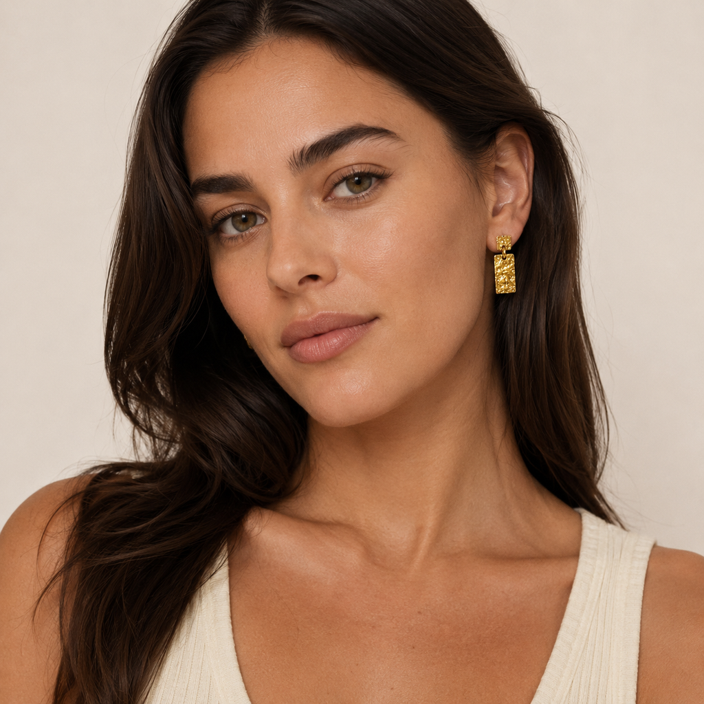 A close-up of a woman wearing a textured gold drop earring, with square top and rectangular drop.