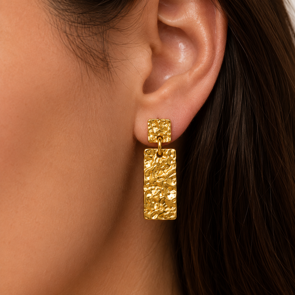 A close-up of a model's ear wearing a textured gold drop earring with a small square stud and a larger rectangular dangling charm.