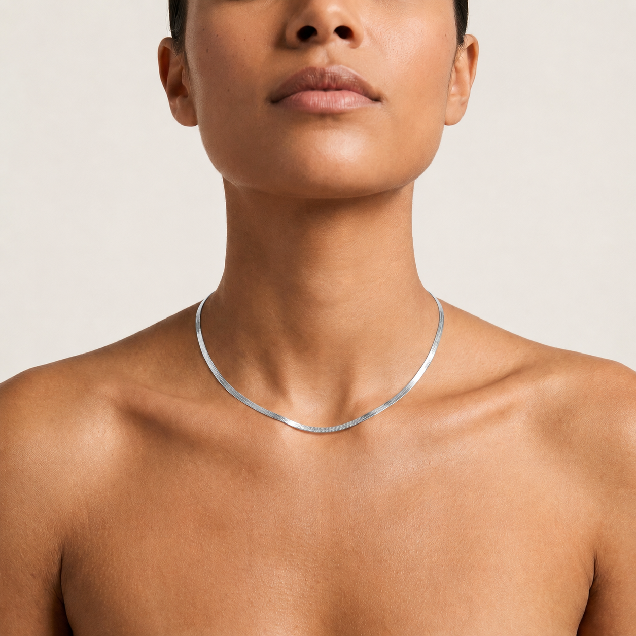 A close-up of a model wearing a thin, polished silver herringbone chain necklace.