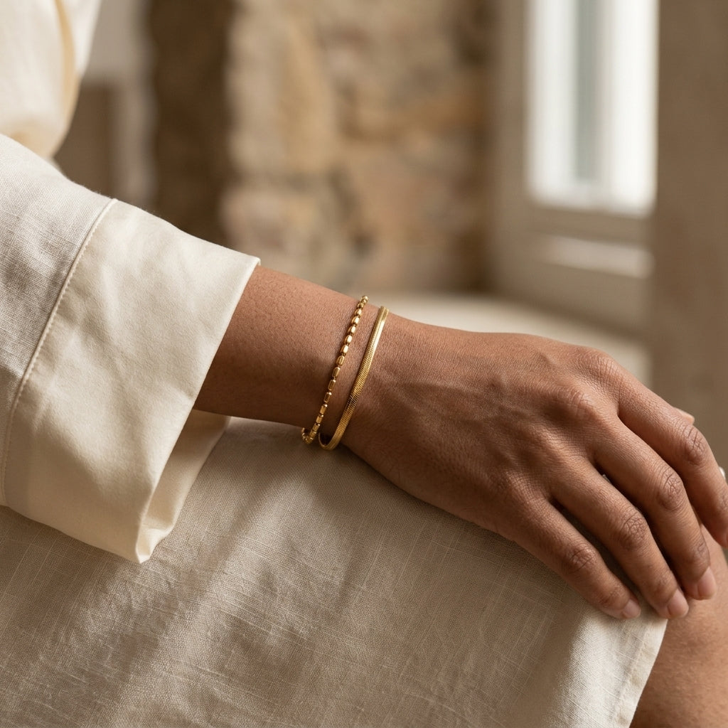 A close-up of a person's hand and wrist wearing a delicate double-layer gold bracelet, one beaded and one herringbone layer.