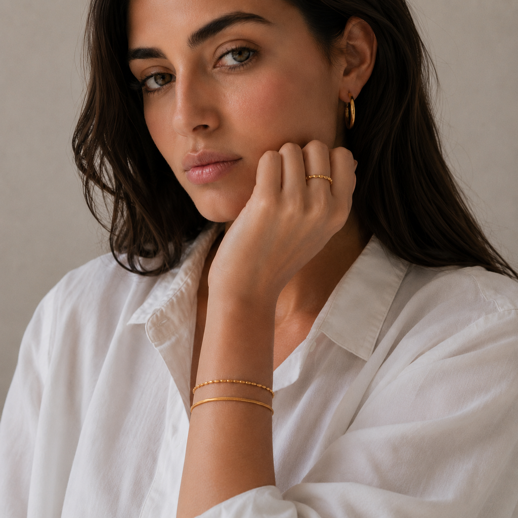 A close-up lifestyle shot of a woman wearing delicate gold jewelry, including a gold beaded bracelet and a gold ring.