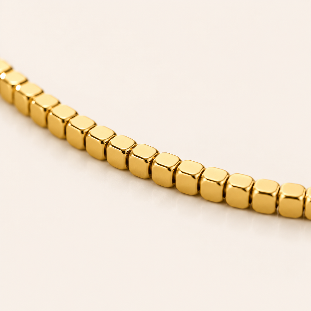 A close-up of a gold-toned beaded anklet with cube-shaped links, photographed on a plain light beige background.