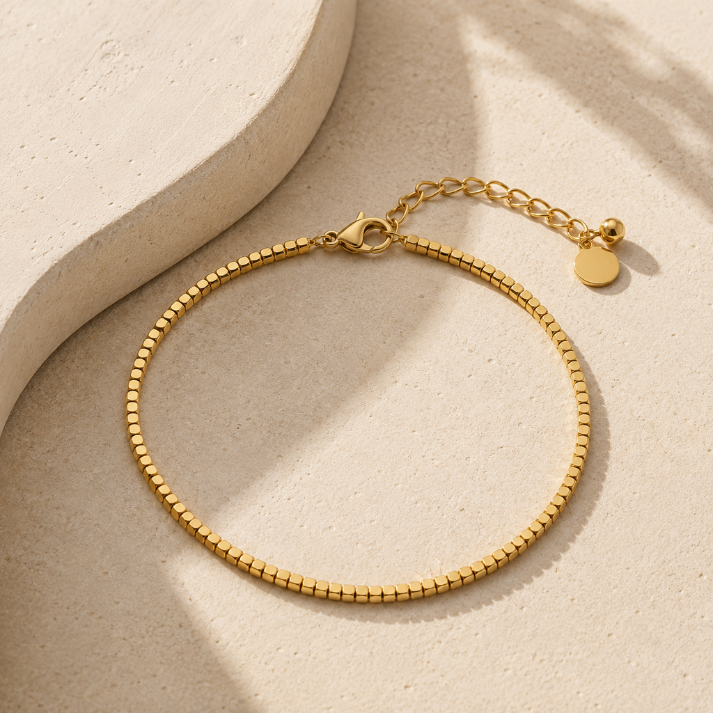 A gold chain anklet with a textured beaded link design, a lobster clasp, an extender chain, displayed on a light beige textured surface with soft shadows.