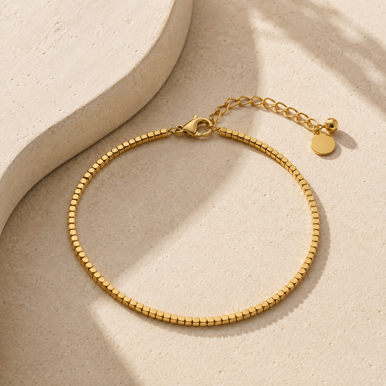 A gold chain anklet with a textured beaded link design, a lobster clasp, an extender chain, displayed on a light beige textured surface with soft shadows.