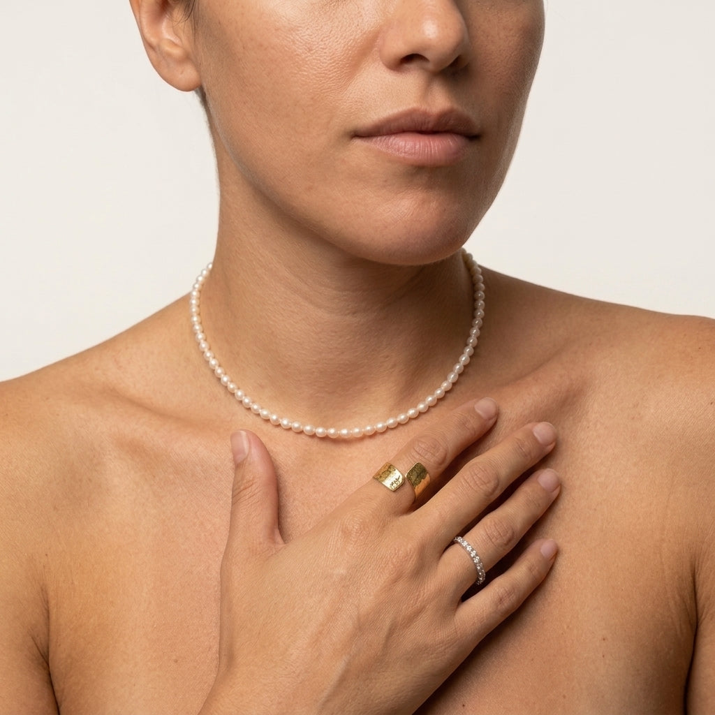 A close-up of a woman wearing a delicate white pearl necklace, with gold rings on her fingers.