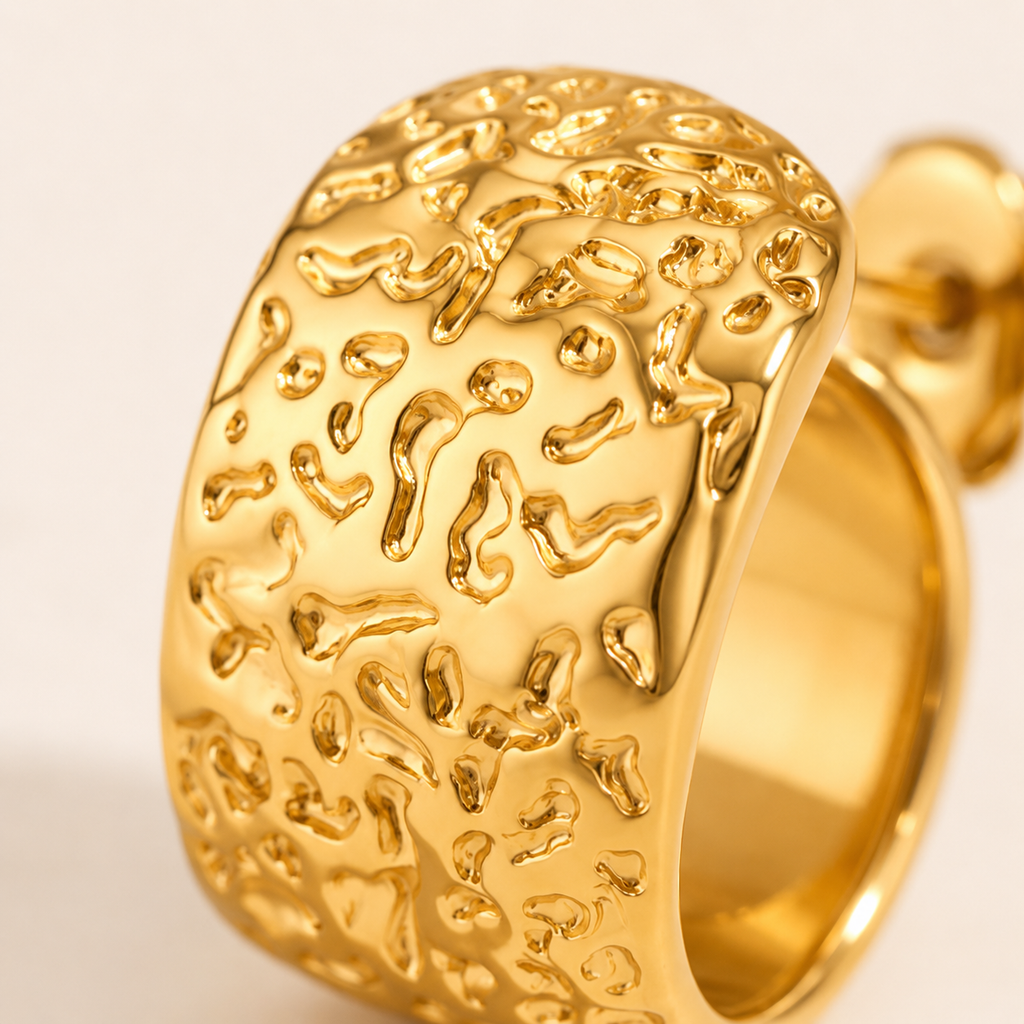 A close-up of a wide, chunky gold earring with a highly textured, organic, hammered surface pattern.