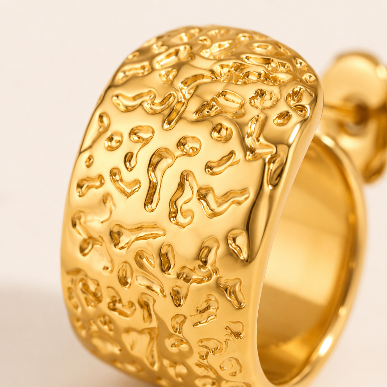 A close-up of a wide, chunky gold earring with a highly textured, organic, hammered surface pattern.