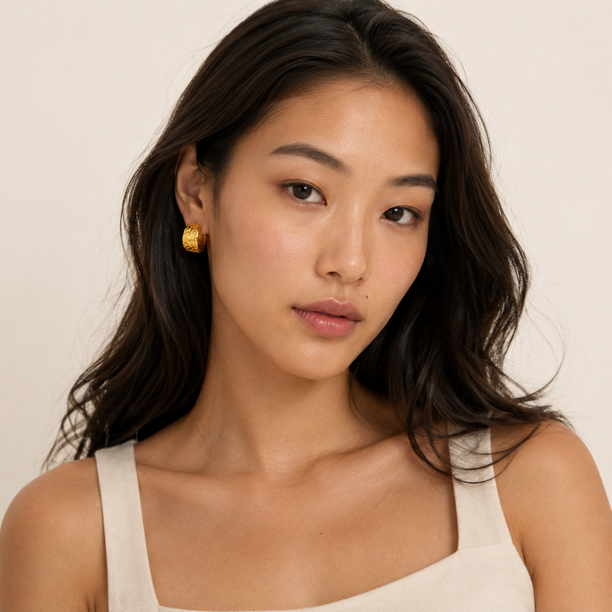 A close-up portrait of a woman with dark hair wearing a textured gold hoop earring.